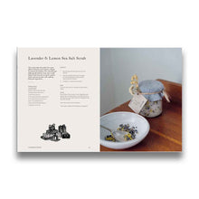 Load image into Gallery viewer, Book page with Lavender & Lemon Sea Salt Scrub with packaging and label on a wooden surface