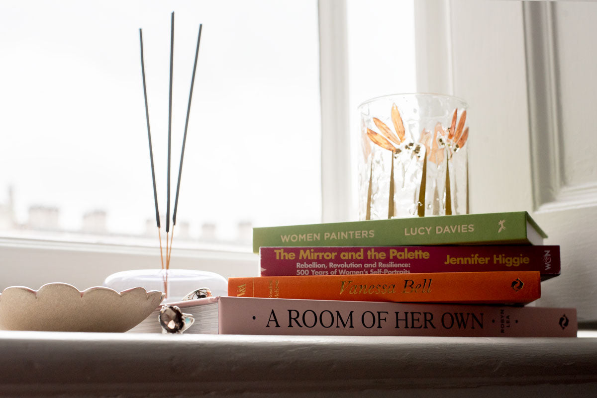 Stack of books written by women on a windowsill with decorative items made by women