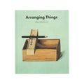 Mint green book titled ‘Arranging Things’ with a minimalist cover design featuring a wooden box, stacked blocks, and a pencil balanced across the top.