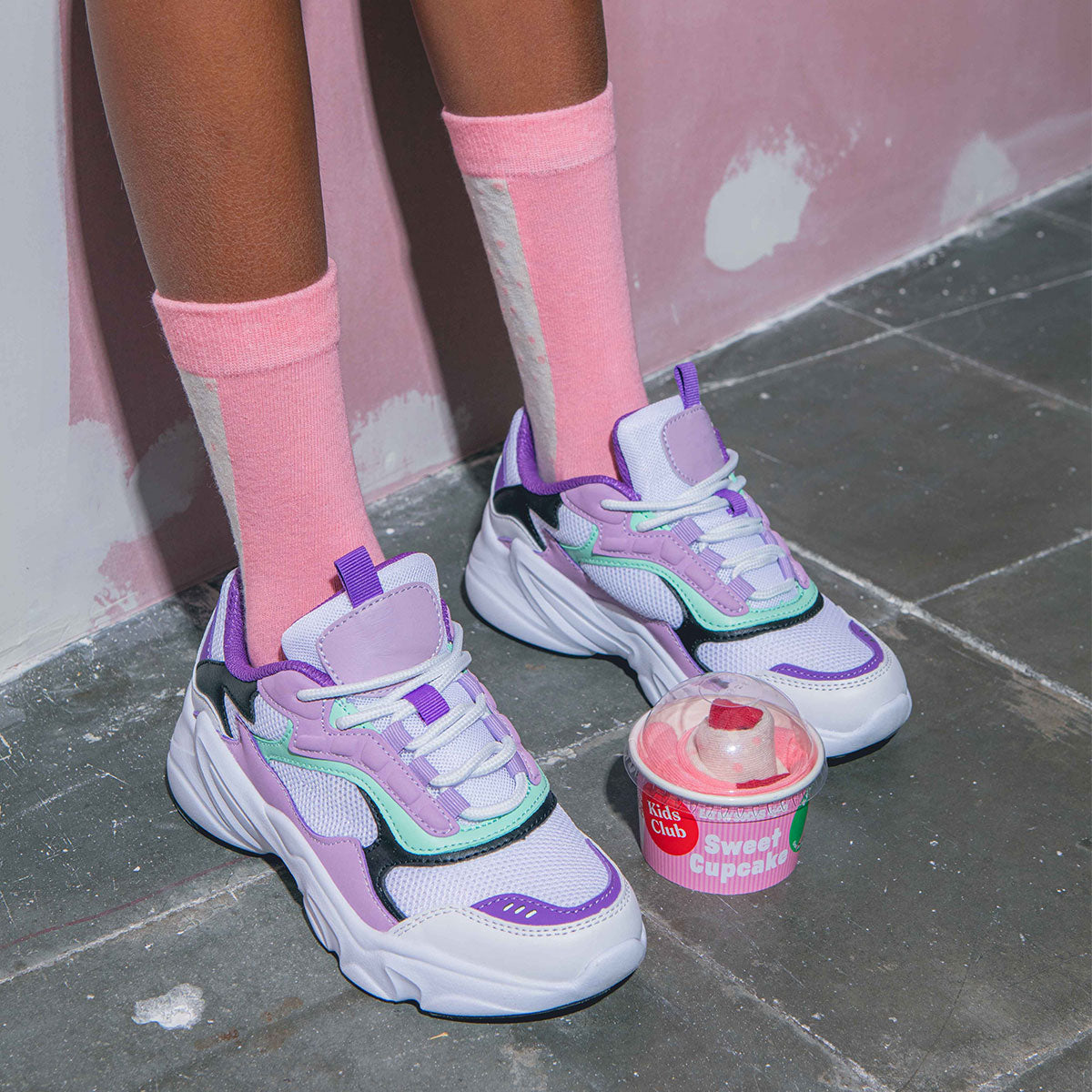 Person wearing colorful sneakers and pink socks on a gray floor with a pink wall.